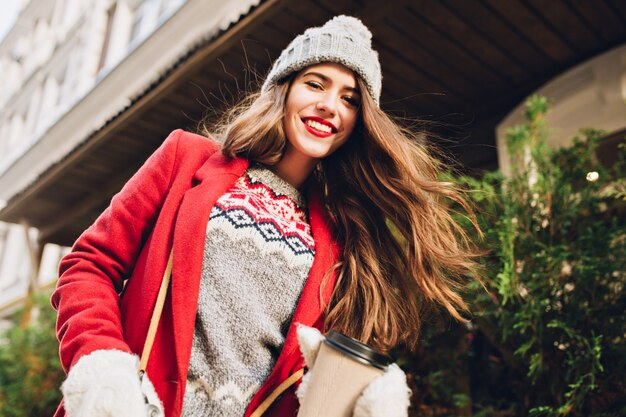 Bella ragazza con i capelli lunghi in cappello lavorato a maglia, cappotto rosso che cammina sulla strada con il caffè per andare. Indossa guanti bianchi, muovendosi dietro la telecamera.