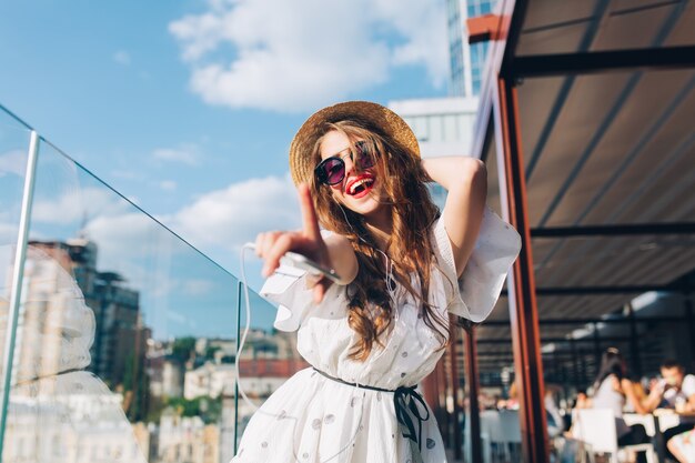 Bella ragazza con i capelli lunghi in occhiali da sole sta ascoltando musica sulla terrazza. Indossa un abito bianco, rossetto rosso e cappello. Sta allungando la mano verso la telecamera e sta ballando. Vista dal basso.