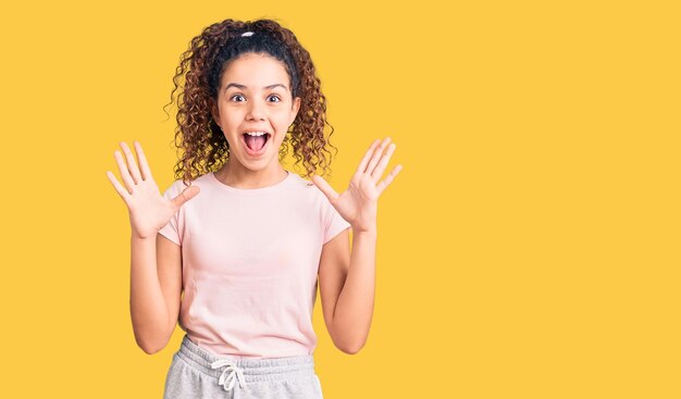 Bella ragazza con i capelli ricci che indossa abiti casual che festeggiano pazzi e stupiti per il successo con le braccia alzate e gli occhi aperti che urlano eccitati. concetto di vincitore