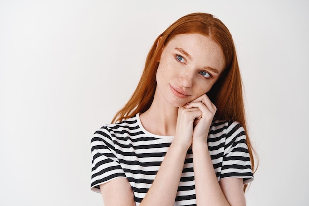 Bella ragazza con i capelli rossi e gli occhi azzurri che guarda con ammirazione e cura la faccia appoggiata della fotocamera sulle mani e guardando a sinistra in piedi su sfondo bianco