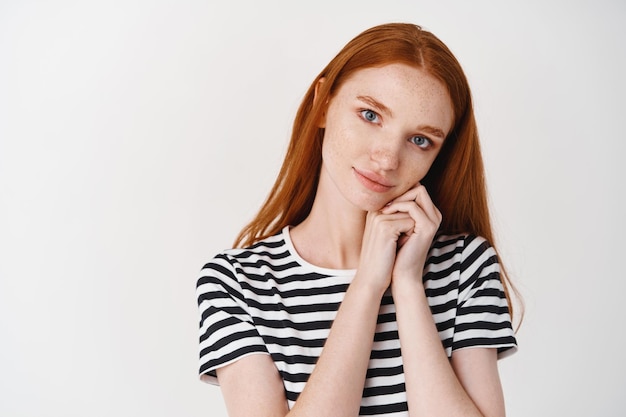 Bella ragazza con i capelli rossi e gli occhi azzurri che guardano con ammirazione e cura davanti, appoggiandosi il viso sulle mani e sognando ad occhi aperti, in piedi sul muro bianco