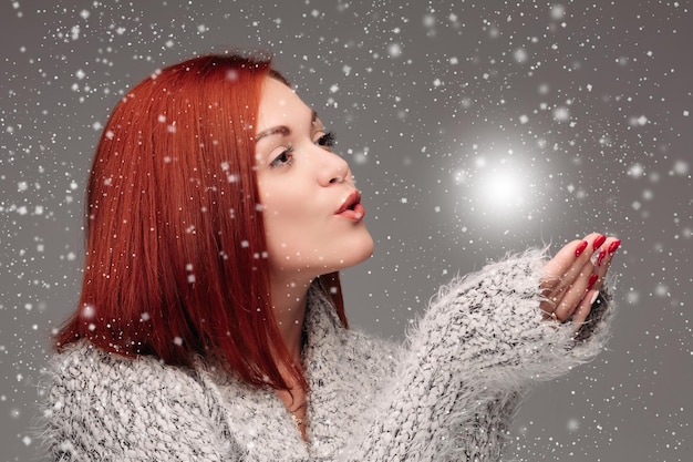 Bella ragazza con i capelli rossi e le unghie rosse che tiene le mani insieme e soffia sulla palla bianca schiarente Bella donna in maglione grigio che cattura la stella e fa i desideri quando nevica