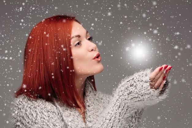 Bella ragazza con i capelli rossi e le unghie rosse che tiene le mani insieme e soffia sulla palla bianca schiarente Bella donna in maglione grigio che cattura la stella e fa i desideri quando nevica