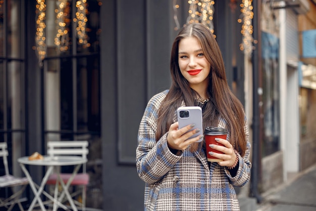 Bella ragazza con i capelli rossi in piedi in un caffè all'aperto e utilizzando un telefono. Ragazza che beve caffè. La ragazza ha le labbra rosse, indossa un elegante cappotto blu.