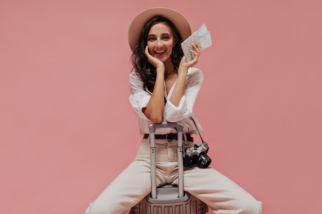 Bella ragazza con i capelli scuri in camicetta leggera e cappello alla moda sorridente e in possesso di due biglietti aerei su uno sfondo isolato