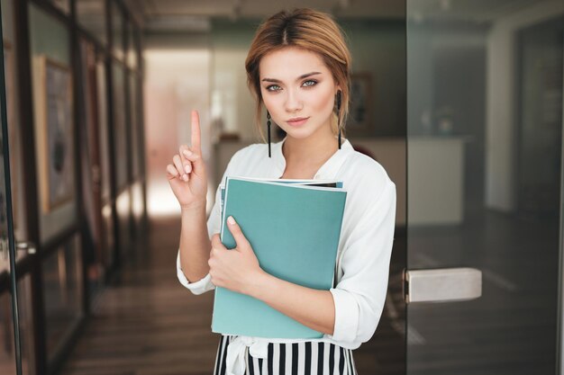 Bella ragazza con i libri in mano che guarda nella fotocamera e non mostra alcun gesto. Ritratto di giovane ragazza con i capelli biondi in camicia bianca in piedi vicino alla porta e con in mano i quaderni.