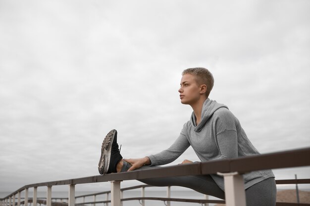 Bella ragazza con il corpo in forma che allunga la gamba usando la guida sulla spiaggia, preparando i muscoli per l'allenamento cardio, avendo un aspetto fiducioso auto determinato Concetto di persone, attività, salute, fitness e sport