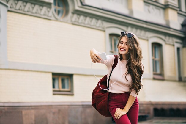 Bella ragazza con labbra vinose e capelli lunghi sta facendo selfie-ritratto in città. Indossa pantaloni vinosi, borsa. Sembra eccitata.