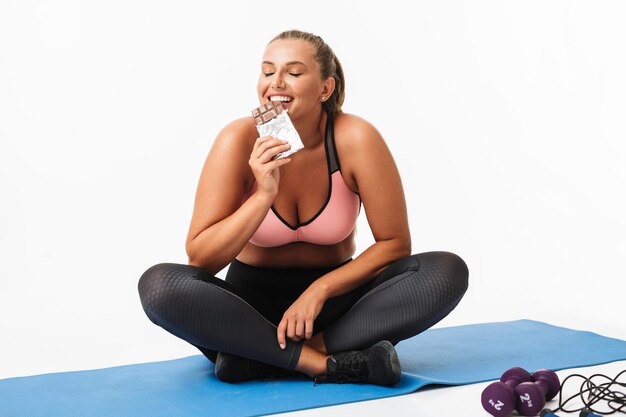Bella ragazza con peso in eccesso in top sportivo e leggings seduti su una stuoia di yoga che mangia felicemente cioccolato su sfondo bianco isolato