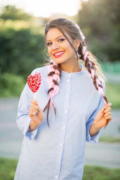 Bella ragazza con trecce colorate in abito blu, tenendo il cuore di caramelle sul bastone.