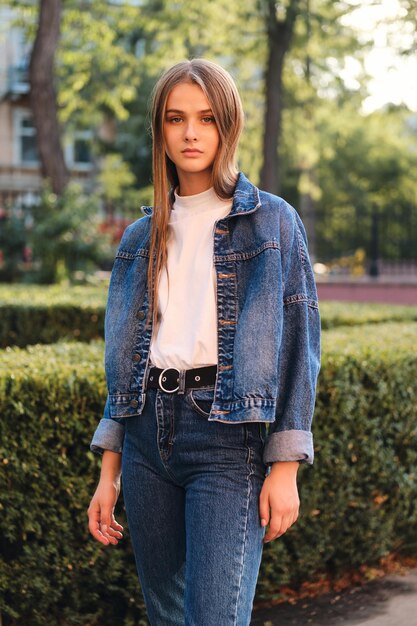 Bella ragazza dai capelli castani casual alla moda in giacca di jeans che guarda seriamente nella fotocamera all'aperto