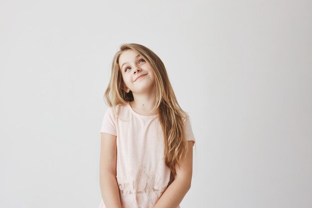 Bella ragazza dai capelli chiari in abito rosa, guardando a testa in su con espressione di viso sexy, pensando di mentire sui segni a scuola per ottenere caramelle dalla madre.