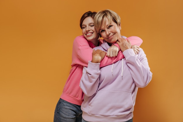 Bella ragazza dai capelli corti in felpa con cappuccio alla moda e jeans che sorride con gli occhi chiusi e che abbraccia sua madre in vestiti alla moda su sfondo arancione.