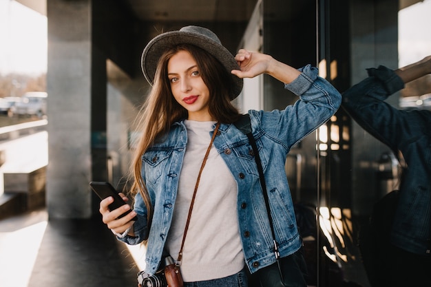 Bella ragazza dai capelli lunghi in vestito di jeans alla moda che cammina fuori e che tiene smartphone nero in attesa di chiamata.