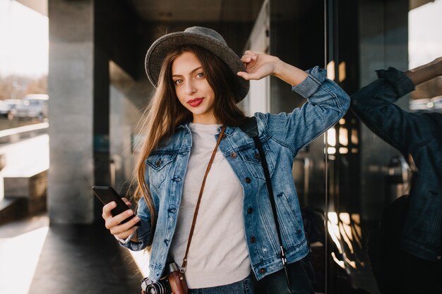 Bella ragazza dai capelli lunghi in vestito di jeans alla moda che cammina fuori e che tiene smartphone nero in attesa di chiamata.