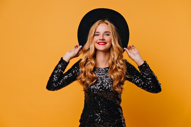 Bella ragazza dai capelli lunghi riccia in posa dopo la festa. Affascinante modello femminile europeo in piedi sul muro giallo.