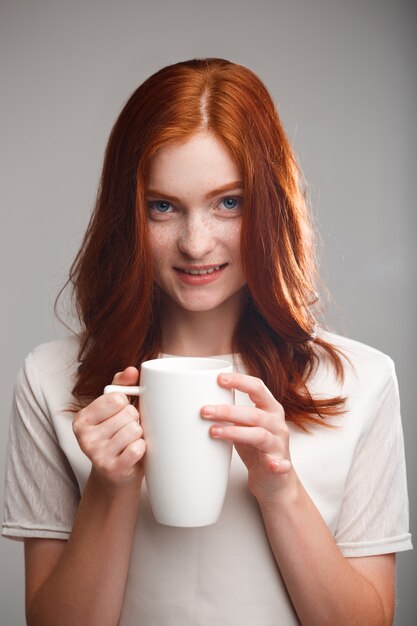 bella ragazza dello zenzero che tiene tazza sopra la parete grigia con luce posteriore.