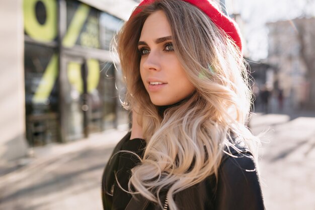 Bella ragazza di moda con capelli biondi lunghi vestito giacca di pelle e cappello rosso cammina per strada alla luce del sole con felici emozioni vere.