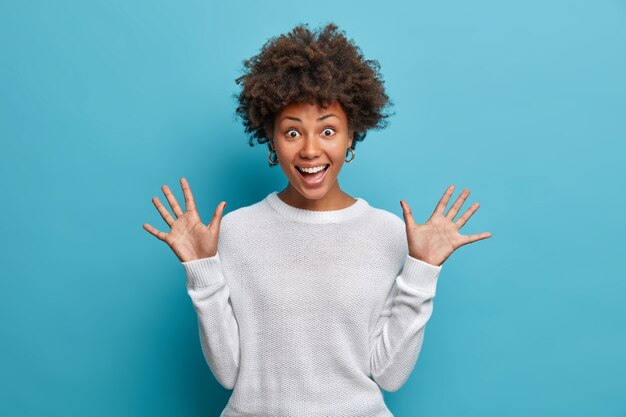 Bella ragazza divertita con i capelli afro ricci, alza i palmi, ha un'espressione allegra, sorride ampiamente, vede qualcosa di divertente, indossa un maglione bianco,