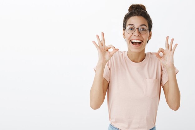 Bella ragazza emozionante con gli occhiali in posa contro il muro bianco