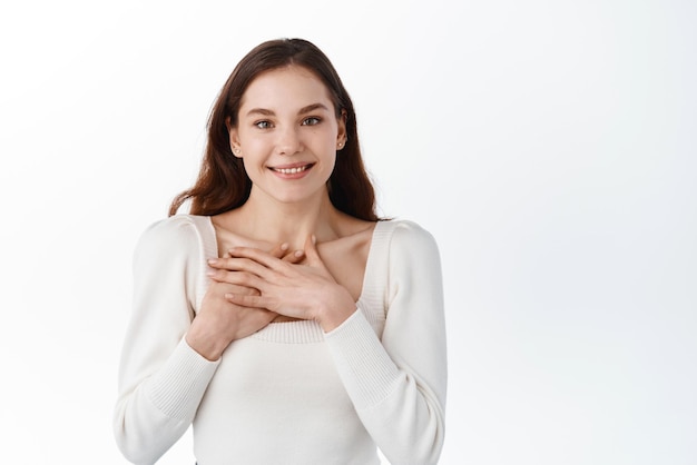 Bella ragazza europea dall'aspetto positivo e amichevole con un bel sorriso sincero che si sente grata e grata mostrando il suo cuore pieno di amore e gratitudine tenendosi per mano sul seno