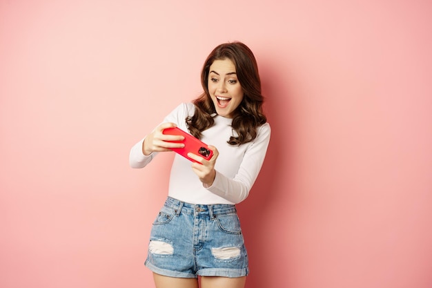 Bella ragazza felice che gioca al videogioco mobile, tenendo lo smartphone in orizzontale, guardando sul cellulare con la faccia eccitata, sfondo rosa.
