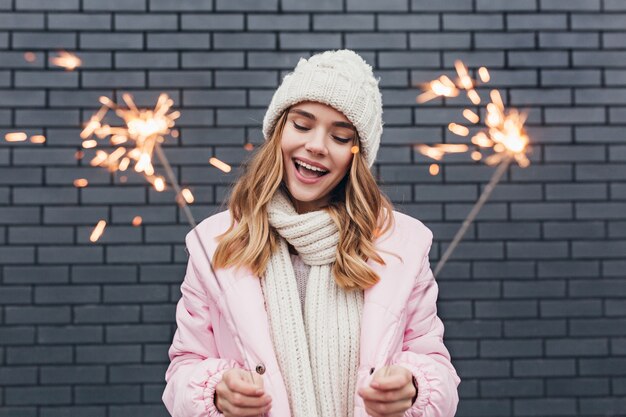 Bella ragazza in abbigliamento invernale bianco che celebra le vacanze. Colpo esterno della signora romantica sorridente che tiene le luci del Bengala.