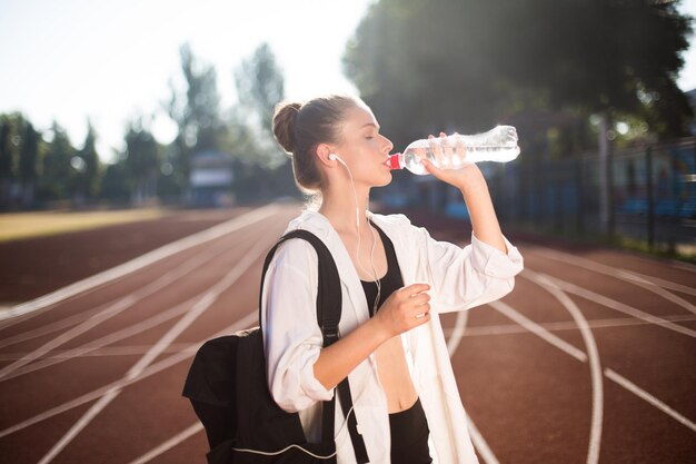 Bella ragazza in abbigliamento sportivo con zaino sulla spalla che beve acqua pura sulla pista dello stadio