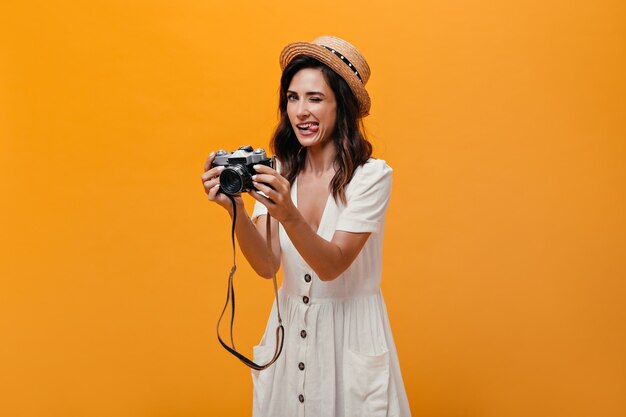 Bella ragazza in abito bianco e cappello mostra la lingua e tiene la fotocamera retrò. Donna divertente in abito leggero estivo con i capelli corti in posa.