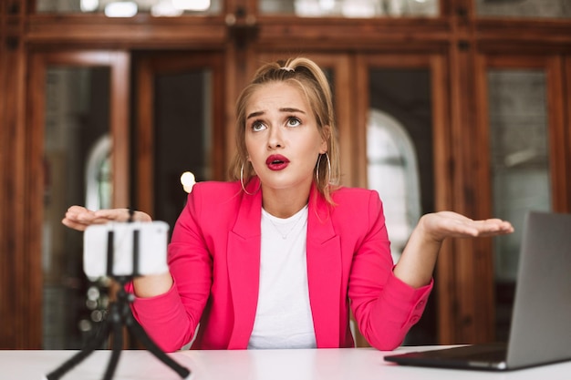 Bella ragazza in giacca rosa che guarda sorprendentemente da parte mentre registra un nuovo video per vlog sul cellulare