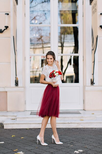 Bella ragazza in gonna in tulle marsala con acconciatura leggera che cammina sulla strada. Tiene i fiori e sorride a lato