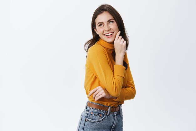 Bella ragazza in piedi mezzo girata e guardando a sinistra con un sorriso allegro, toccando la guancia e fissando felice, indossando abiti casual su bianco