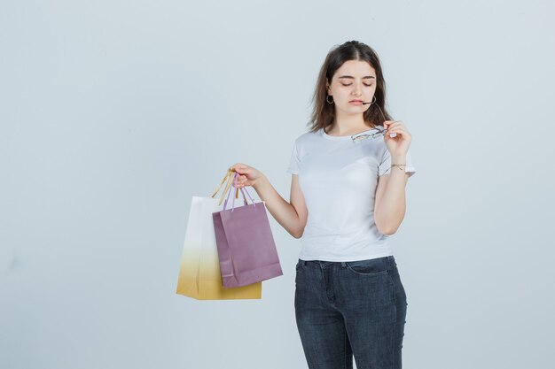 Bella ragazza in t-shirt, jeans mantenendo sacchetti regalo e occhiali e guardando triste, vista frontale.