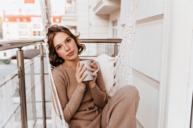 Bella ragazza in vestito lavorato a maglia che beve caffè al mattino. Giovane donna caucasica romantica che tiene tazza di tè al balcone.