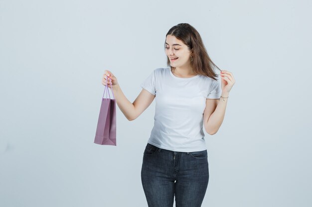Bella ragazza mantenendo la borsa papper in t-shirt, jeans e guardando felice, vista frontale.