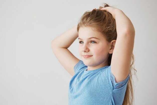 Bella ragazza minuscola con affascinanti occhi azzurri e lunghi capelli chiari sorrisi, guardando da parte e facendo acconciatura. Concetto di infanzia, infanzia e stile di vita.