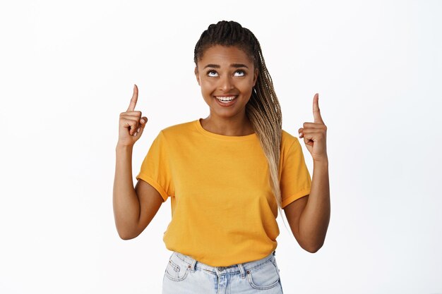Bella ragazza nera sorridente che punta il dito verso l'alto guardando l'offerta promozionale di vendita speciale in piedi su sfondo bianco