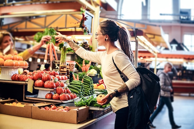 Bella ragazza pensierosa con gli occhiali sta comprando carote fresche al mercato agricolo locale.