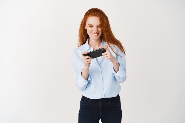 Bella ragazza rossa che gioca al videogioco su smartphone e sorride. Donna inclina il telefono cellulare e sembra gioiosa, in piedi su un muro bianco