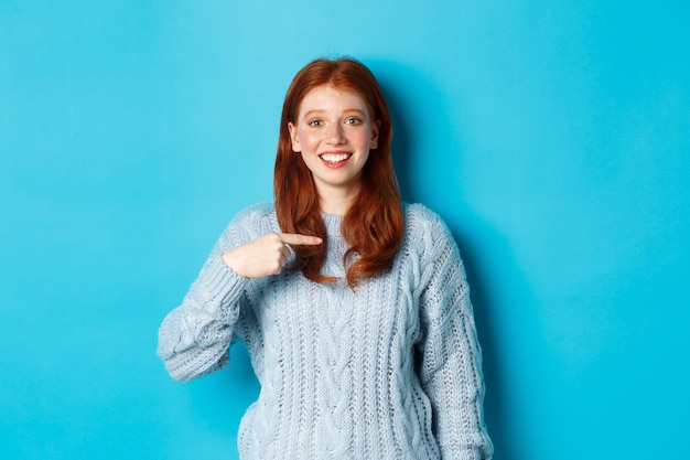 Bella ragazza rossa che indica se stessa e sorride felice, essendo scelta, in piedi in maglione su sfondo blu.