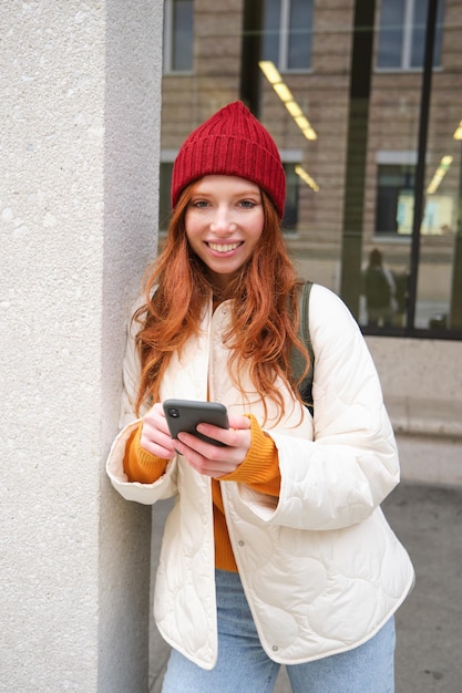 Bella ragazza rossa con smartphone in piedi sulla strada che tiene il telefono cellulare utilizzando l'app taxi connec