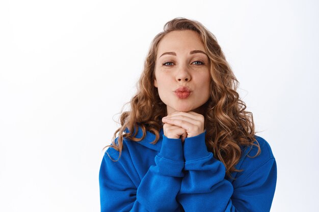 Bella ragazza sciocca con capelli biondi ricci, labbra arricciate, che bacia la faccia e simpatica espressione facciale, in piedi civettuola sul muro bianco