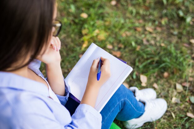 Bella ragazza seria dai capelli scuri in giacca di jeans e occhiali fa i compiti nel parco verde estivo.