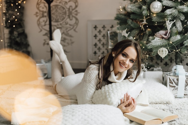 Bella ragazza si trova con cuscini e plaid vicino a un albero di Natale