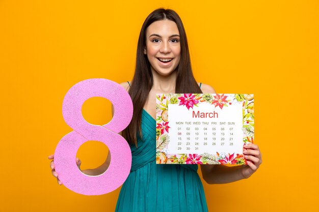 Bella ragazza sorpresa il giorno della donna felice che tiene il calendario con il numero otto isolato sulla parete arancione