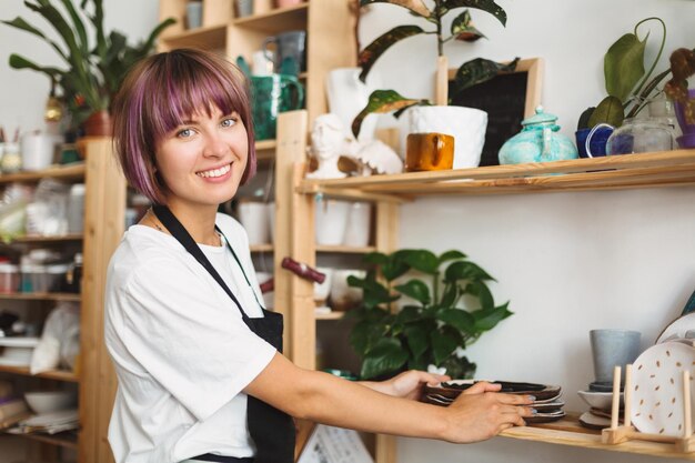 Bella ragazza sorridente con capelli colorati in grembiule nero e maglietta bianca che mette piatti fatti a mano sullo scaffale guardando con gioia nella fotocamera allo studio di ceramica