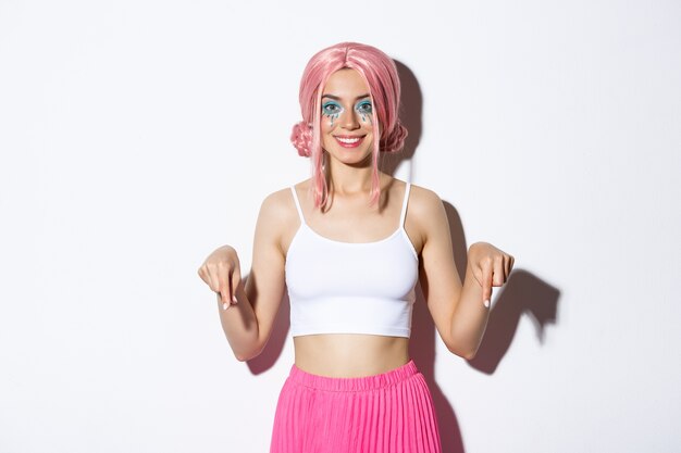 Bella ragazza sorridente con i capelli rosa e trucco luminoso, puntando il dito verso il tuo logo, indossando il costume di Halloween, in piedi.