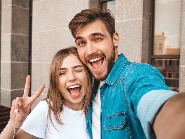 Bella ragazza sorridente e il suo ragazzo bello in abiti casual estivi. Famiglia felice che prende selfie autoritratto di se stessi sulla fotocamera dello smartphone. Mostra il segno di pace e ammiccanti in strada