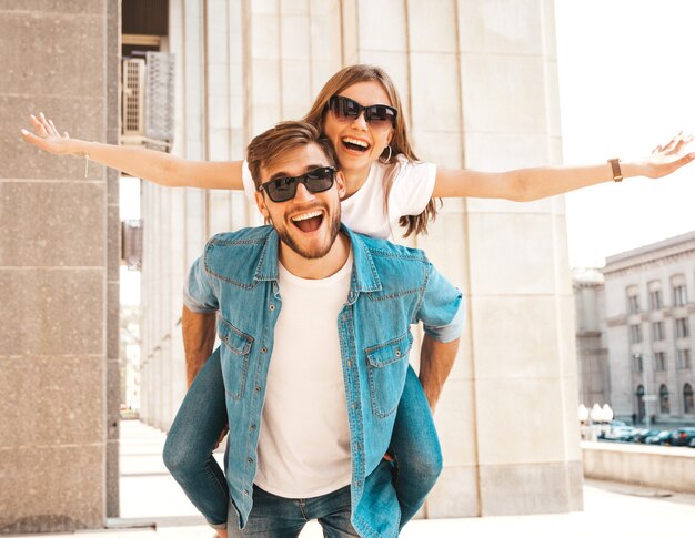 Bella ragazza sorridente e il suo ragazzo bello in abiti casual estivi. Uomo che porta la sua ragazza sulla schiena e lei alzando le mani.