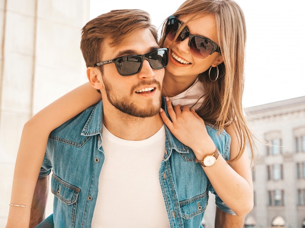 Bella ragazza sorridente e il suo ragazzo bello in abiti casual estivi. Uomo che porta la sua ragazza sulla schiena e lei alzando le mani.